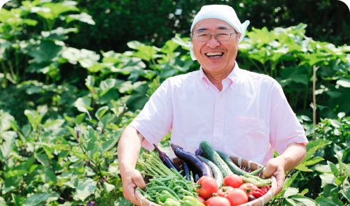 爽緑農園の太田紘一さんが、カゴに入ったきゅうりや茄子などの野菜を笑顔で見せてくれている写真